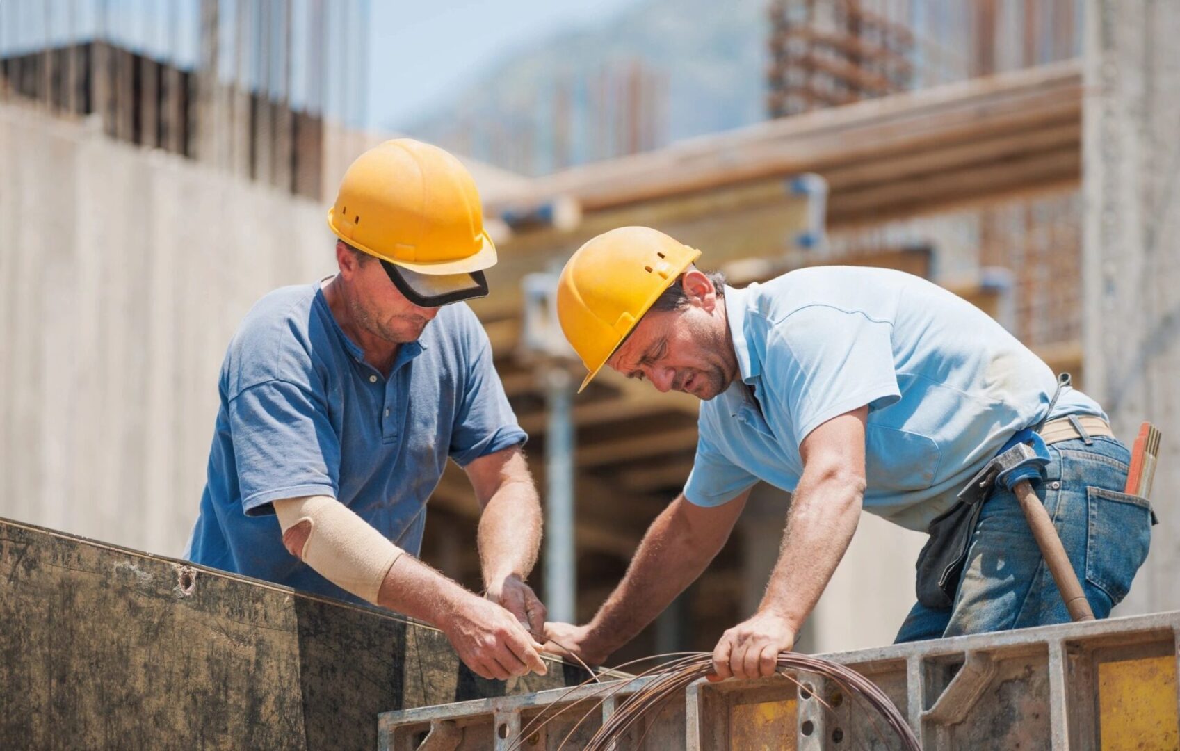 Two construction workers wearing yellow hard hats are diligently working with wires at a construction site. One worker holds a bundle of wires while the other uses a tool, showcasing their expertise in Professional Building Services. Building structures stand prominently in the background.